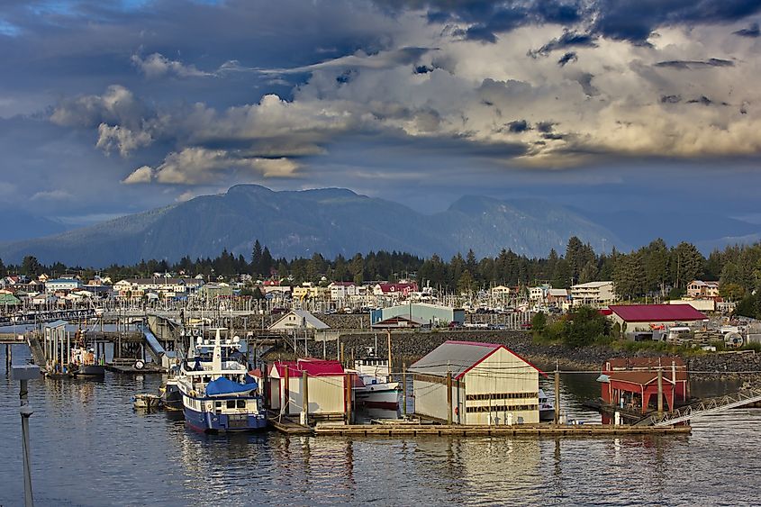 Scenic harbor view with boats docked alongside colorful, red-roofed buildings. In the background, mountains rise under a dramatic, cloudy sky.