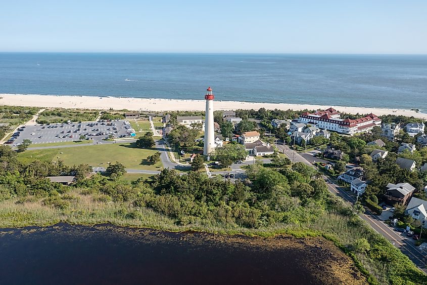 Aerial view of a coastal town with a prominent white lighthouse with a red top, surrounded by greenery. Nearby, a beach borders the blue ocean. Houses and a parking area are visible, conveying a serene, summery vibe.