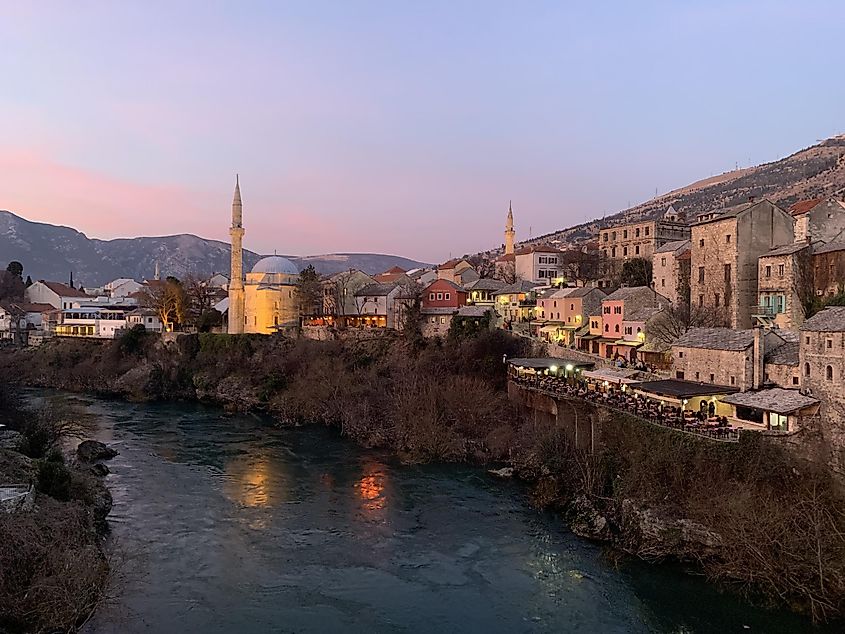 Evening descends on Mostar. Mosques and restaurants can be seen lining the Neretva River beneath a purple sky.