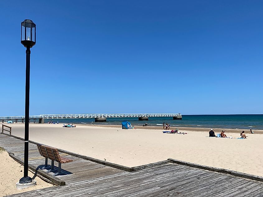 A boardwalk with lamp post runs parallel to a sandy beach and perpendicular to a distant pier.