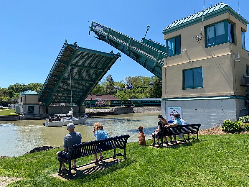 A small sailboat passes under a raised drawbridge while onlookers watch from riverside benches.