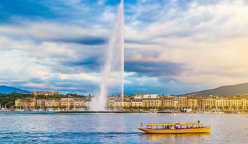 Panoramic view of Geneva skyline with famous Jet d'Eau fountain and traditional boat at harbor district in beautiful evening light at sunset, Canton of Geneva, Switzerland