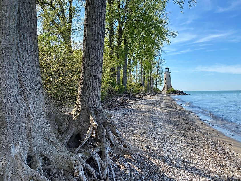An old stone lighthouse stands at the end of a slim, forested beach.