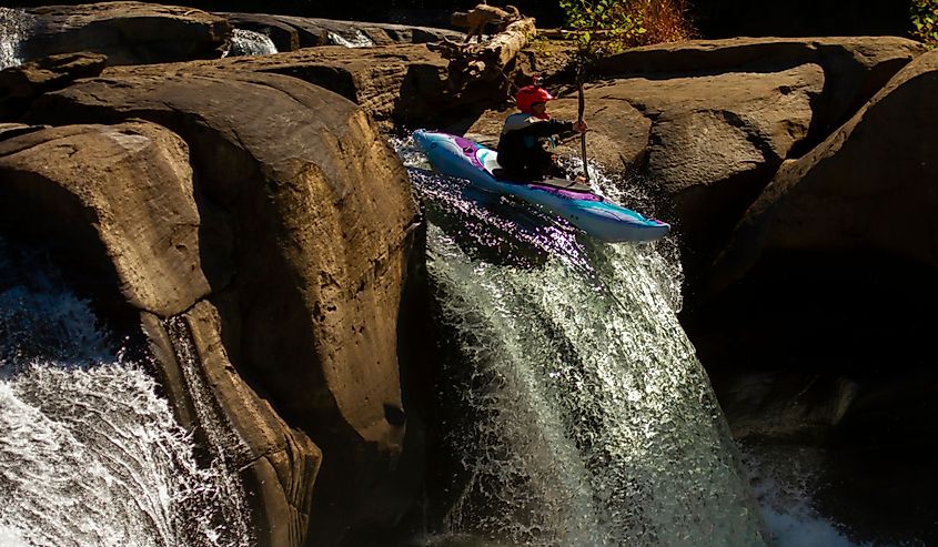 A kayaker wearing a red helmet with camera attached to it is doing a brave performance by descending down the fast flowing rocky waterfall in Ohiopyle Falls.
