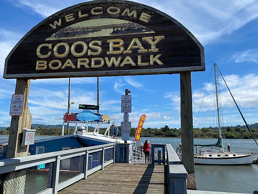 A woman in a red jacket walks onto a dock, beneath a large sign for "Coos Bay Boardwalk" and toward a riverside seafood restaurant. 