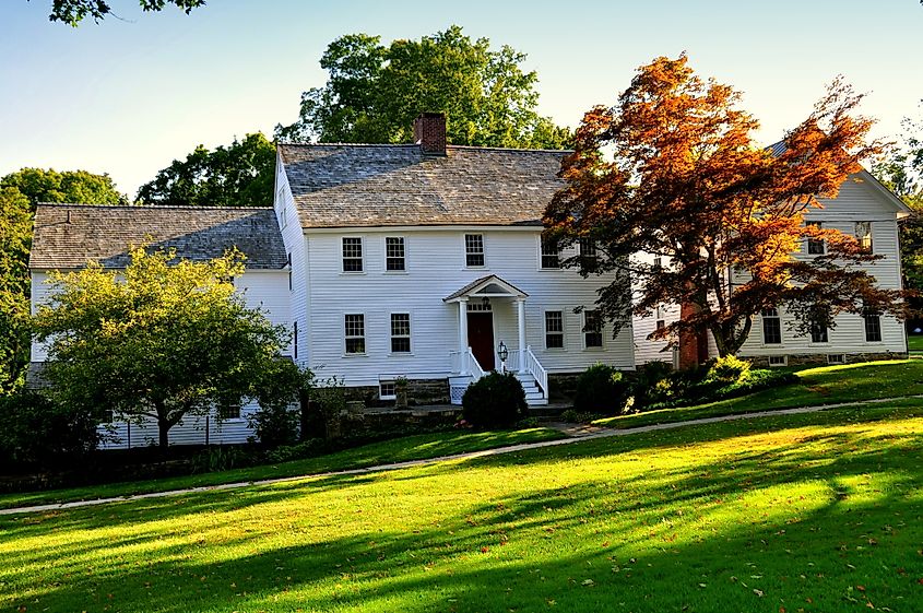 White colonial-style house in Washington, Connecticut with a red front door, surrounded by green lawn, trees, and fall-colored leaves in warm sunlight.