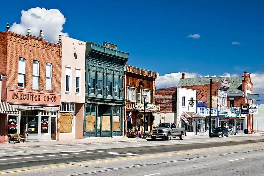 Historic small-town street with rustic storefronts, brick buildings, and vintage signage under a clear blue sky. A few vehicles are parked alongside.