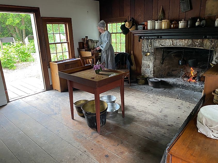 A period actor in early 1800s attire prepares a meal in an old fashioned kitchen using a wood fire.