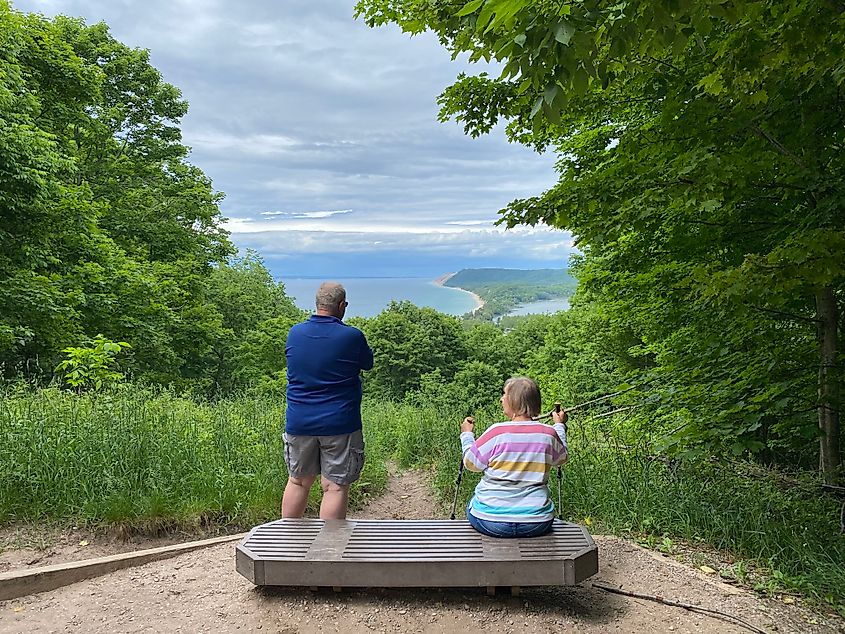 An elderly couple pauses on a bluff bench to take in the lake, forest, and sand dune views.