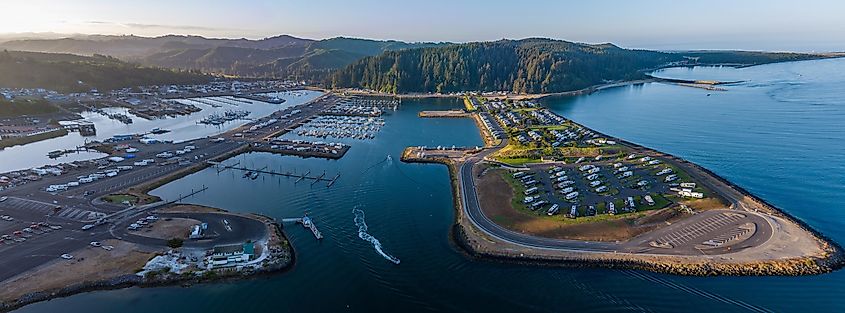 Aerial view of Winchester Bay, Oregon, featuring the residential district, harbor, marina, a large RV park, and fishing boats heading out in the calm water at sunrise.