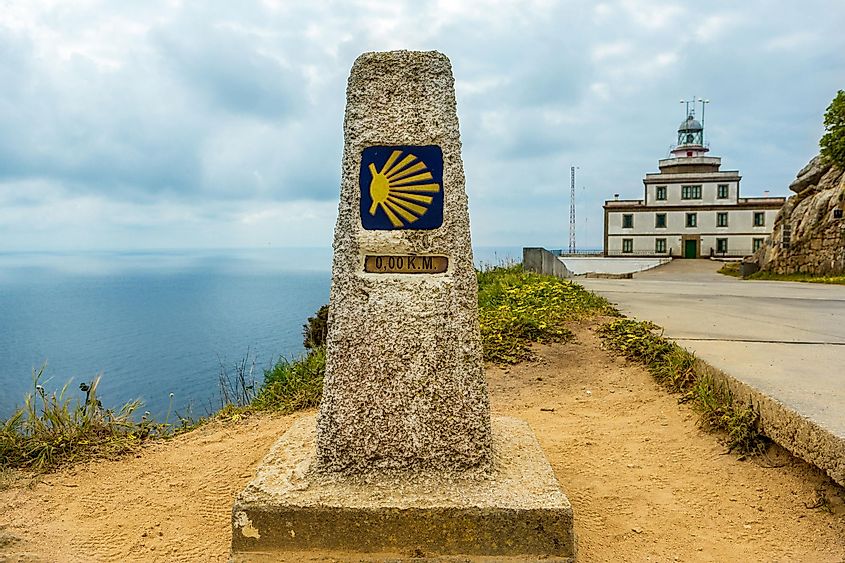 The emblematic Camino de Santiago kilometer-marker shows 0.00 at Cape Finisterre. A lighthouse can be seen on the right, and the blue expanse of the Atlantic Ocean to the left.  