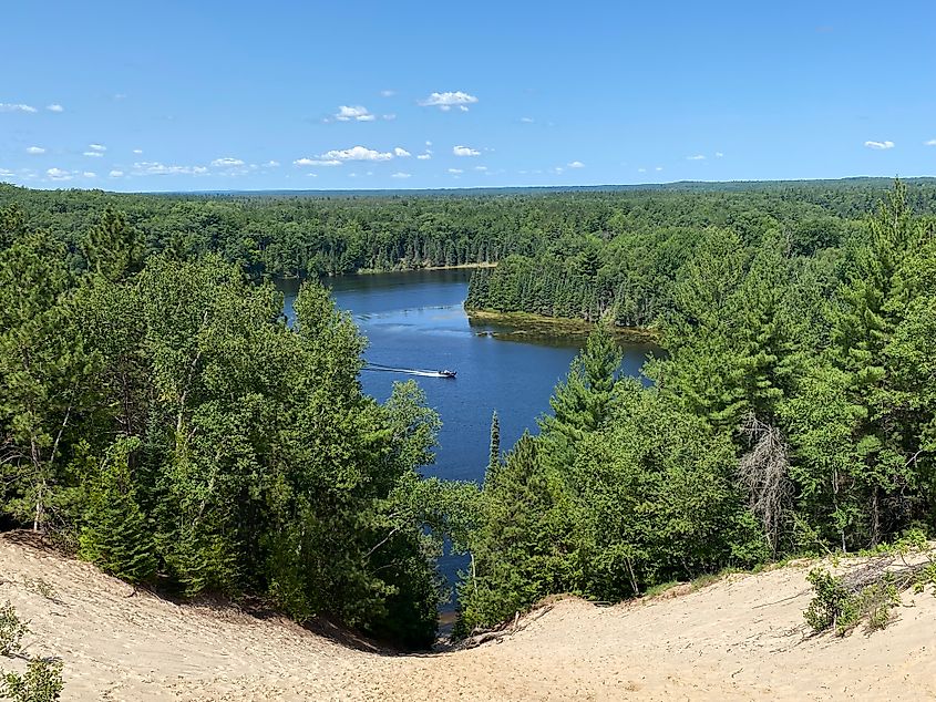 A motor boat seen from high above cruising around a river bend, through an immense forest, past a giant sand dune.