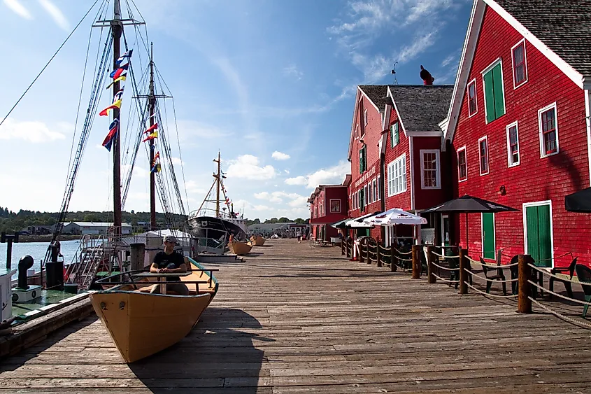 A sunny seaside dock featuring vibrant red buildings with green shutters, colorful flags on sailboats, and a person sitting in a small wooden boat.