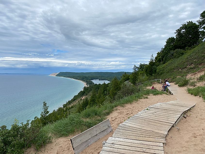 Hikers admire the viewpoint at the end of a sandy, forest trail overlooking a long, Lake Michigan shoreline.