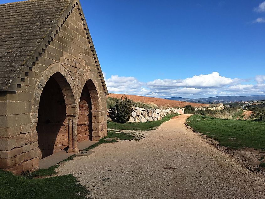 A stone monastery stands beside a dirt road, leading off into the hilly Spanish countryside on a blue sky day.  