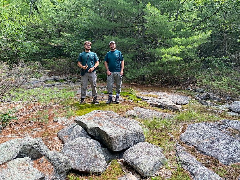 Two young men in green, national park employee shirts pose for a picture in a rocky forest.