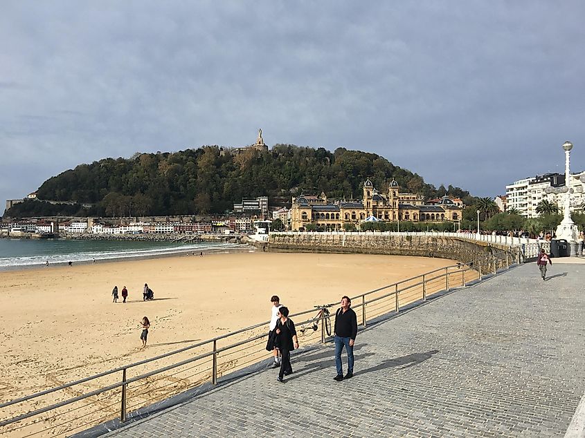 People walk a beachside boardwalk in front of a hillside Spanish old town.
