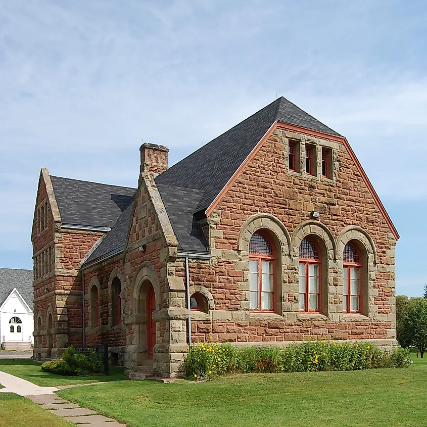 Historic stone building with arched windows and a steep, dark roof. The structure is surrounded by a green lawn and garden under a clear blue sky.