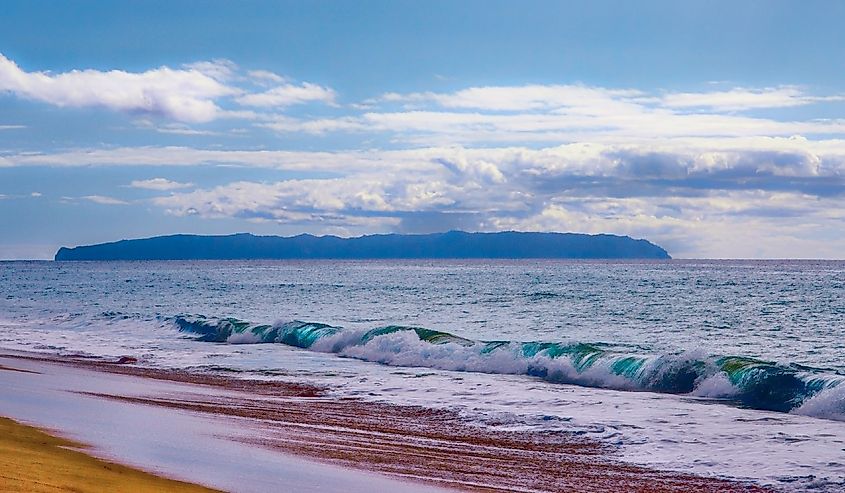 Distant view of Niihau with the crashing waves on a sandy beach in the forefront