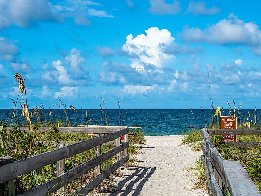 Sandy walkway with wooden railings leading through sea oats to a beach at Stump Pass State Park in Englewood, Florida, with the Gulf of Mexico and a partly cloudy sky ahead.