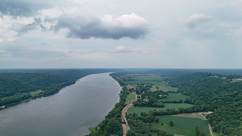A wide aerial view shows the Ohio River curving through the landscape near Madison, Indiana. On one side of the river are green fields, scattered homes, and a winding road, while dense forest covers the opposite bank. Overhead, a cloudy sky casts soft light across the rural scenery.