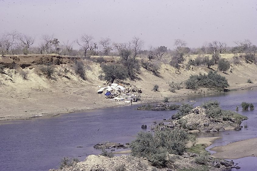 Two men are drying washed clothes on the bank of the Falémé River at a low water level in a dry Sahelian landscape of low bushes and low trees on the banks