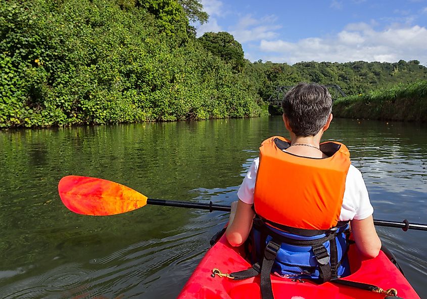 A retired caucasian lady in a red canoe on Hanalei River approaching the old steel bridge in Kauai