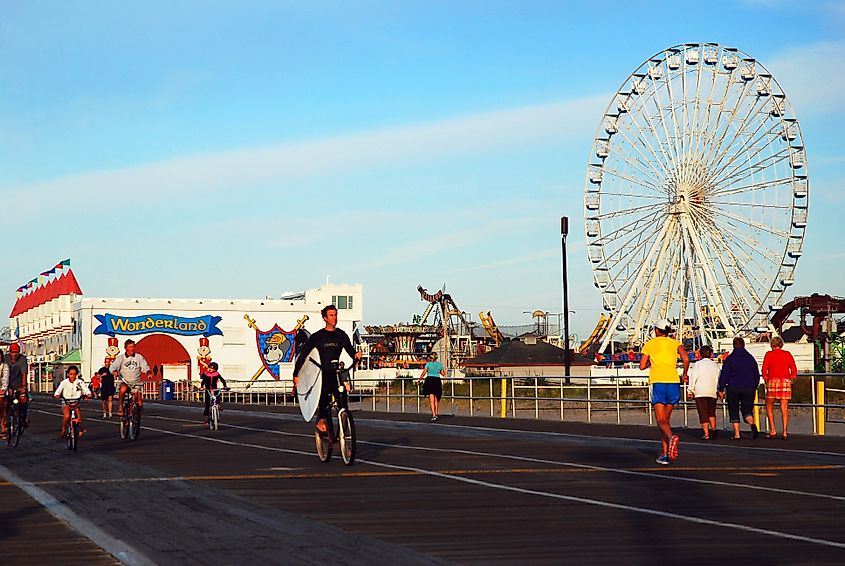 People walk, jog, and ride bikes along the Ocean City, New Jersey boardwalk beside amusement rides, including a large Ferris wheel, under a clear blue sky.