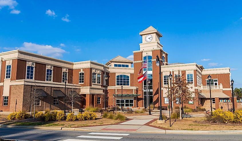 Street view of Lilburn City Hall, showing outside sitting area, main entrance doors and windows, and pole with flags of USA and Georgia, under fair skies