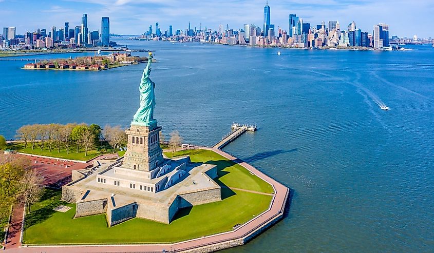 Aerial view of the Statue of Liberty on a sunny day, surrounded by water. The New York City skyline is visible in the distance, evoking grandeur.
