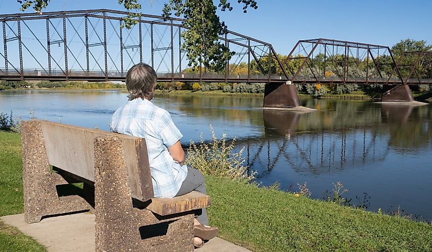 Senior woman sitting on a bench admiring the historic Fort Benton Bridge spanning the Missouri River in Montana.
