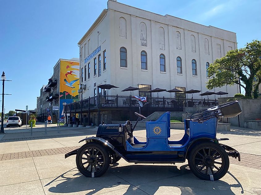 An antique Ford car branded "Sandusky" parked on the sidewalk of a colorful, well-kept downtown street.