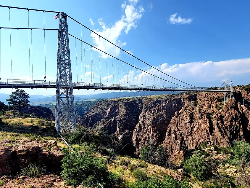 Suspension bridge crossing a deep rocky canyon in Cañon City, Colorado, with steep cliffs, sparse vegetation, and a wide blue sky in the background.