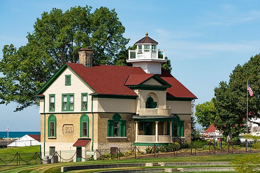 A historic lighthouse with a red roof and green trim stands amid lush greenery under a clear sky. The scene exudes a serene, timeless charm.