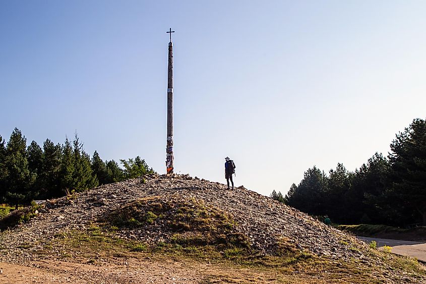 A male hiker stands atop a mountain of stones, looking up at a tall pole with a crucifix at its top. 