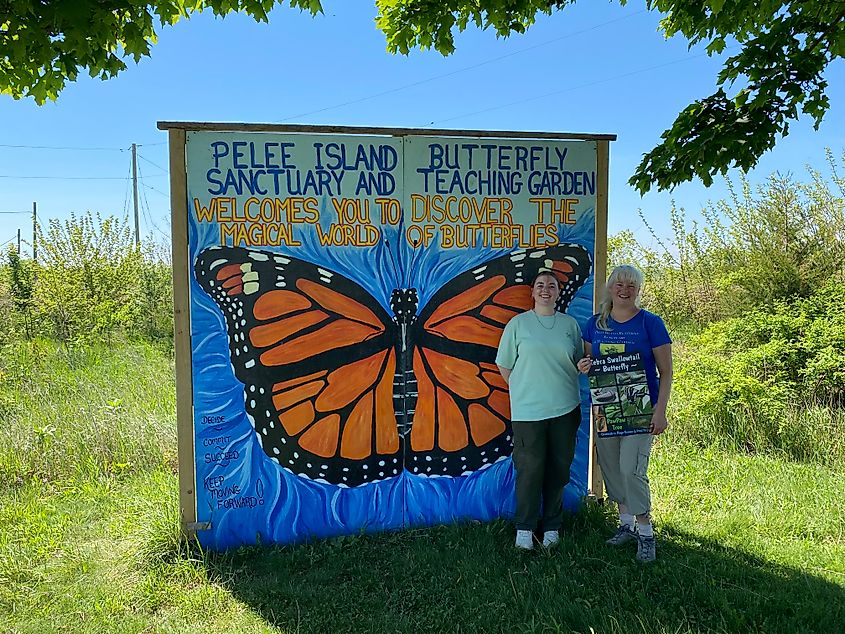 Two women pose in front of a large mural of a monarch butterfly advertising the Pelee Island Butterfly Sanctuary.