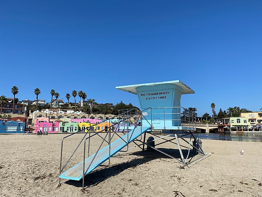 A blue lifeguard tower stands in the middle of a sandy beach with brightly colored lodgings in the background.