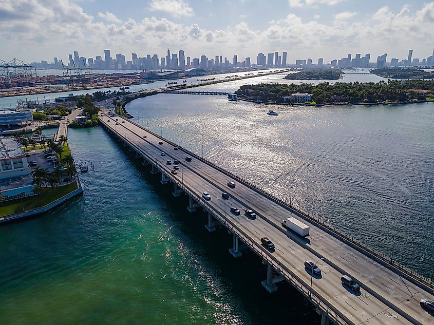 Aerial view of a coastal highway bridge in Florida with cars traveling over blue-green water toward a distant city skyline