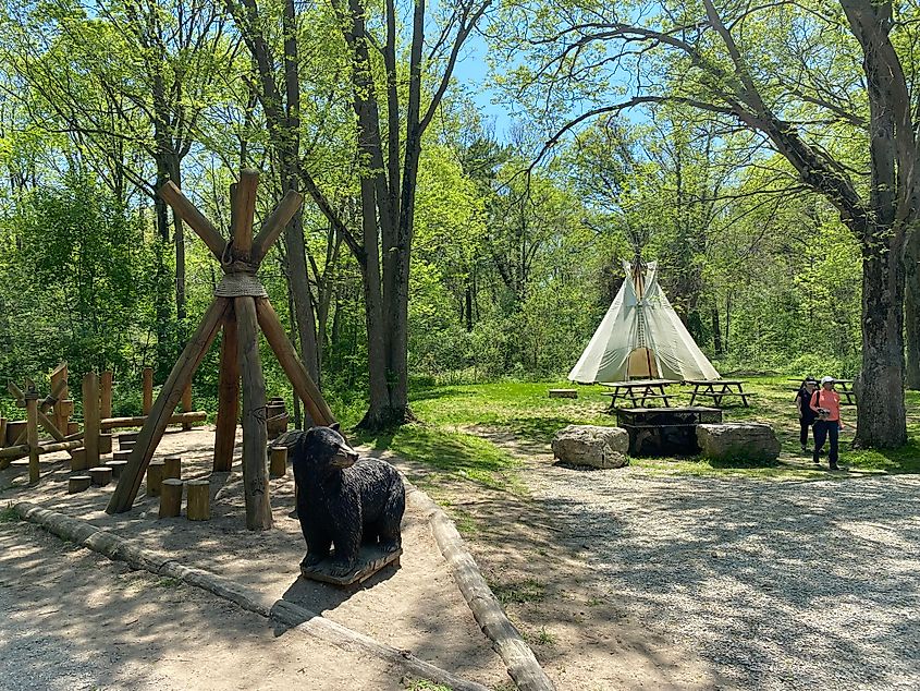 A teepee is set up in the grass behind a wooden playground with a black bear statue.