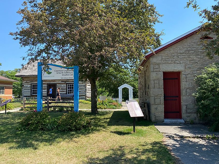 A couple investigates a log cabin. The town sign indicates that the stone building beside it is North America's Smallest Jail.