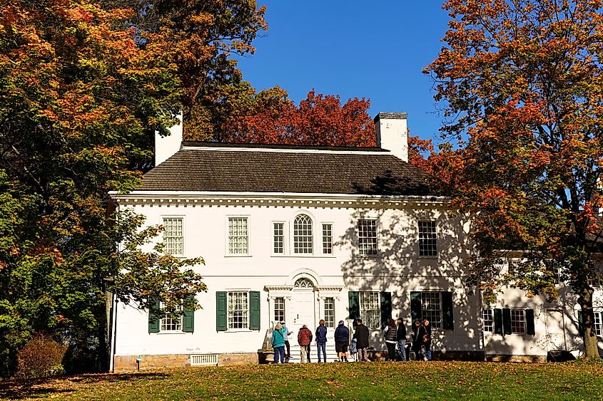 Historic white house with green shutters surrounded by vibrant autumn trees. A group of people stands in front, enjoying the sunny day.