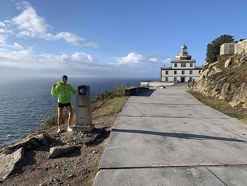 A young woman poses next to a trail marker, in front of a lighthouse at the tip of a Spanish cape. The sun shines on the North Atlantic Ocean