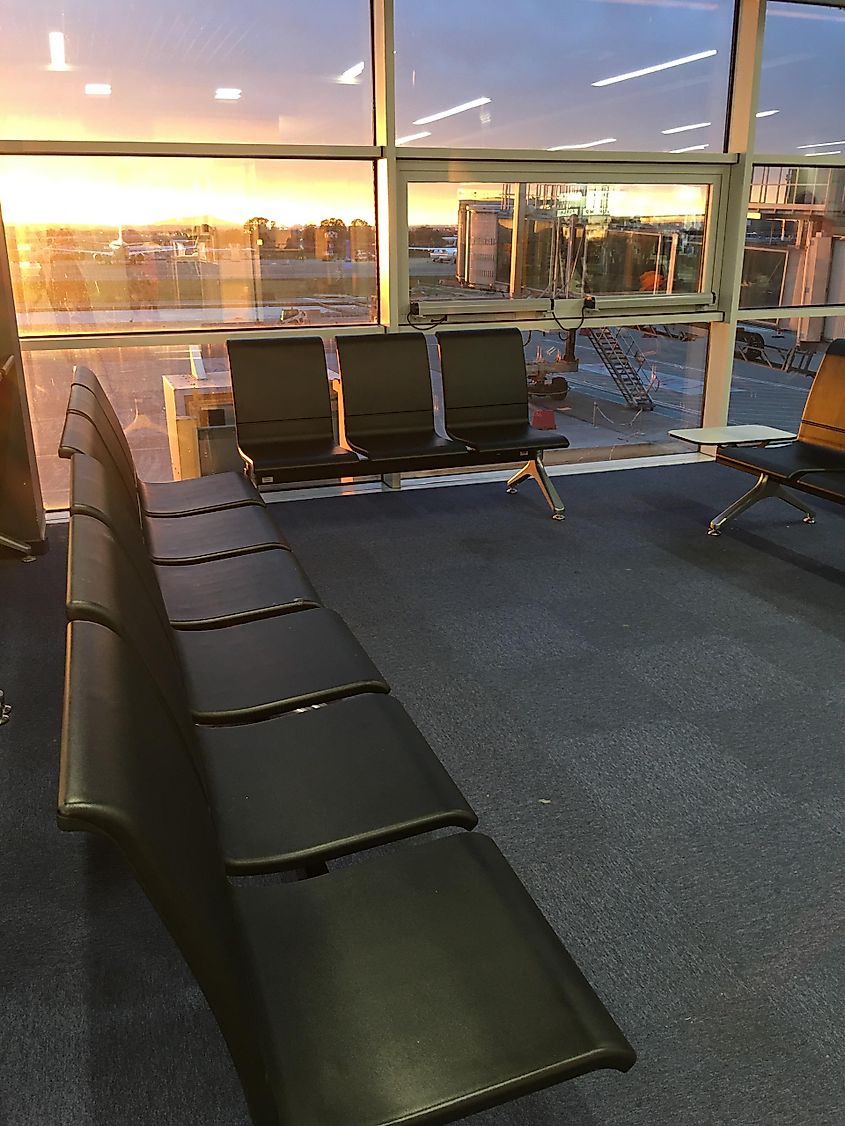 A row of empty black airport chairs in an empty terminal. The sun rises over an airport setting.  
