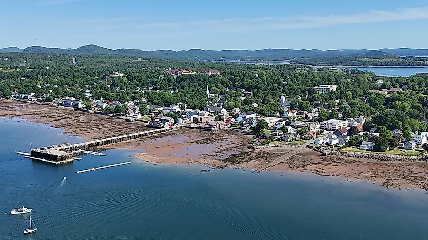 Aerial view of a quaint coastal town with a long pier stretching into calm waters, surrounded by lush green hills and scattered white houses under a clear blue sky.