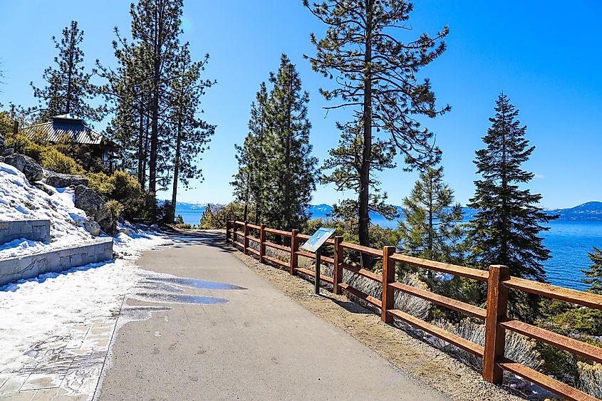 A long smooth concrete walking path with a wooden fence near vast deep blue lake water with lush green trees and plants with snow and blue sky at Lake Tahoe Nevada State Park in Incline Village Nevada.