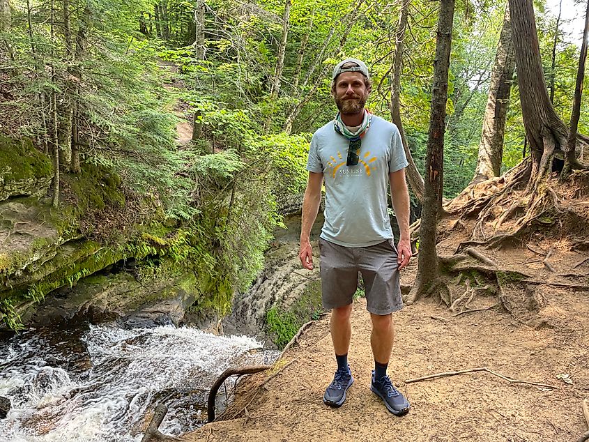 A male hiker poses at the top of a wooded waterfall.