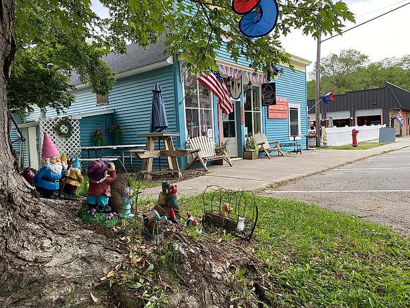 A collection of garden gnomes sits at the base of a tree before a blue ice cream shop.