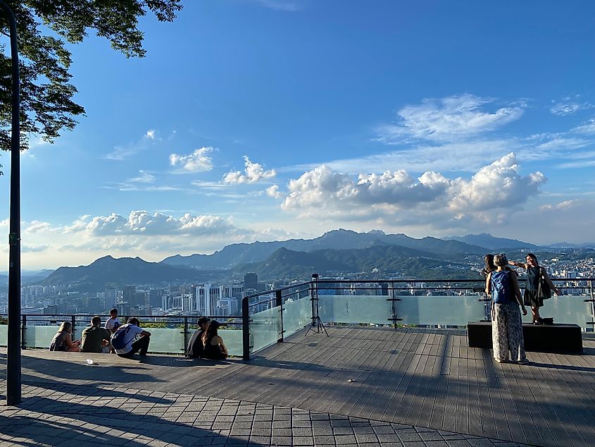 A small crowd gathers and poses for pictures atop a mountain top viewing platform overlooking Seoul, South Korea.