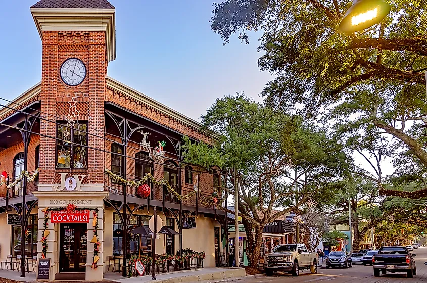 A two-story red-brick building with a clock tower stands on a corner in Ocean Springs, Mississippi. The building is decorated with holiday garlands and ornaments, including a large 'JOY' sign and festive lights. A restaurant occupies the ground floor, with outdoor seating along the sidewalk. Large oak trees draped with string lights line the street, where several cars are parked.