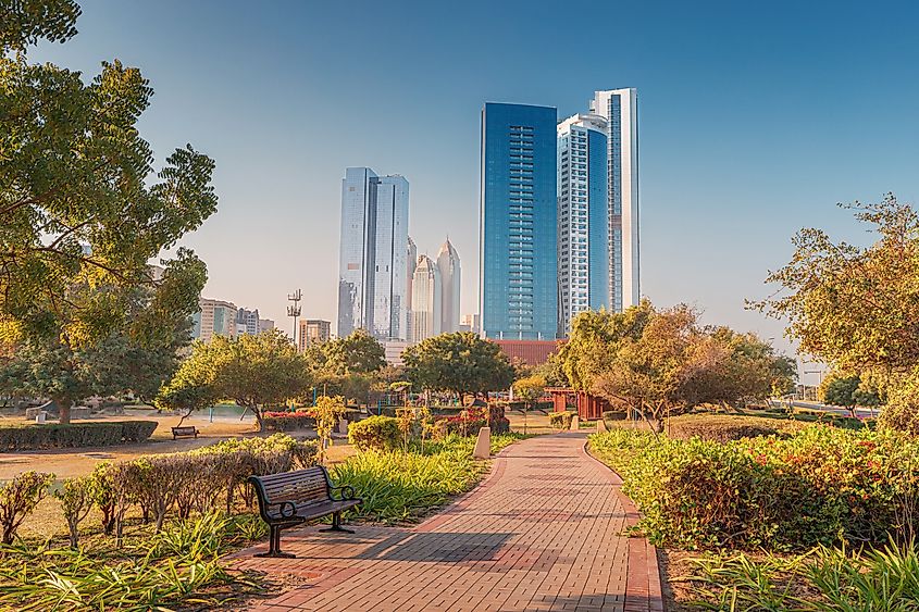 Empty park with bench and brick path leading to modern skyscrapers in Abu Dhabi during sunny morning.